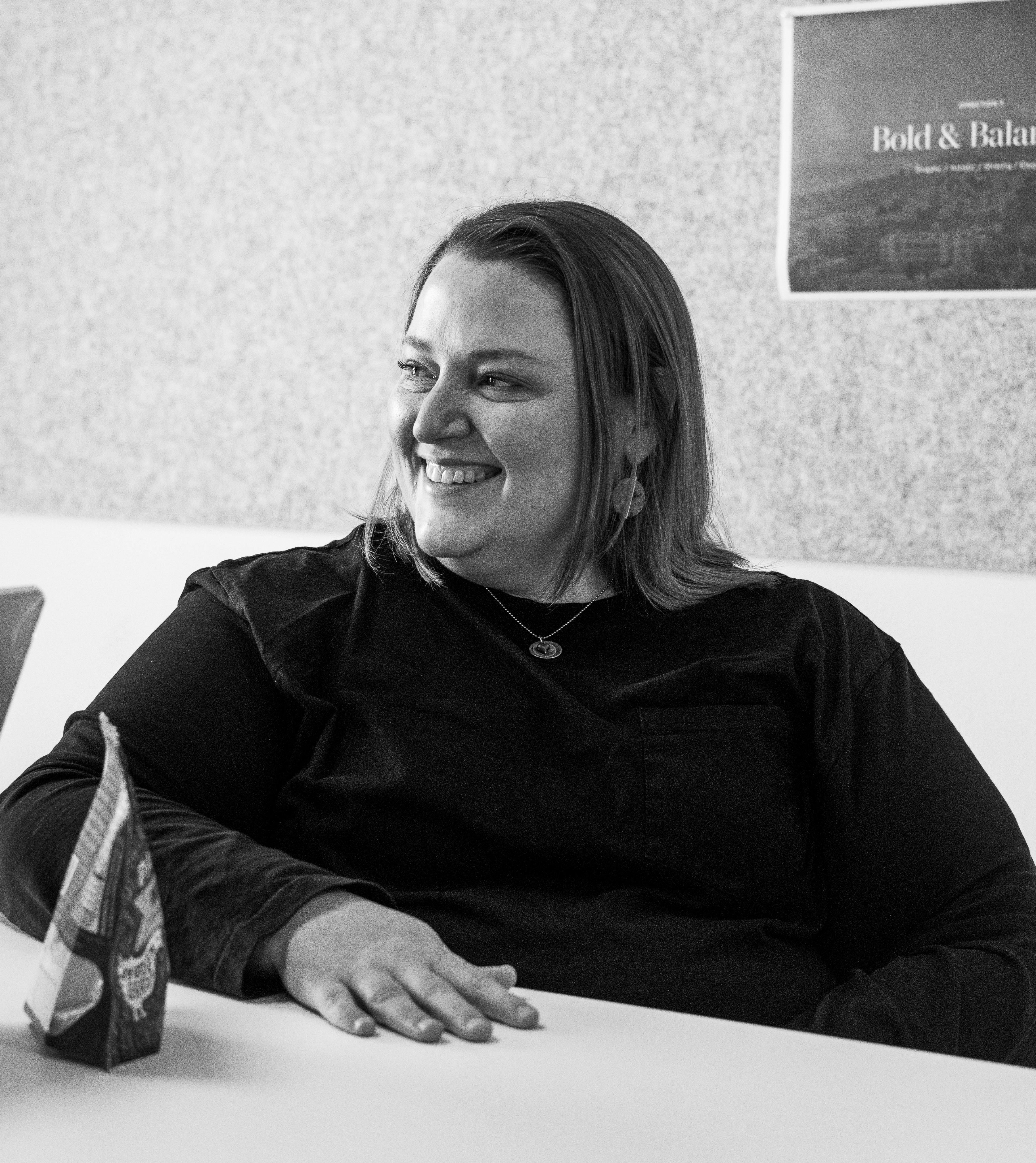 Business Duos A smiling woman in a dark shirt sits at a table, exuding the confidence often associated with married couples in business and entrepreneur couples. Behind her, a partially visible poster on the wall provides subtle context. This black-and-white image highlights the professional poise common to husband-and-wife business partners who successfully combine marriage and business.