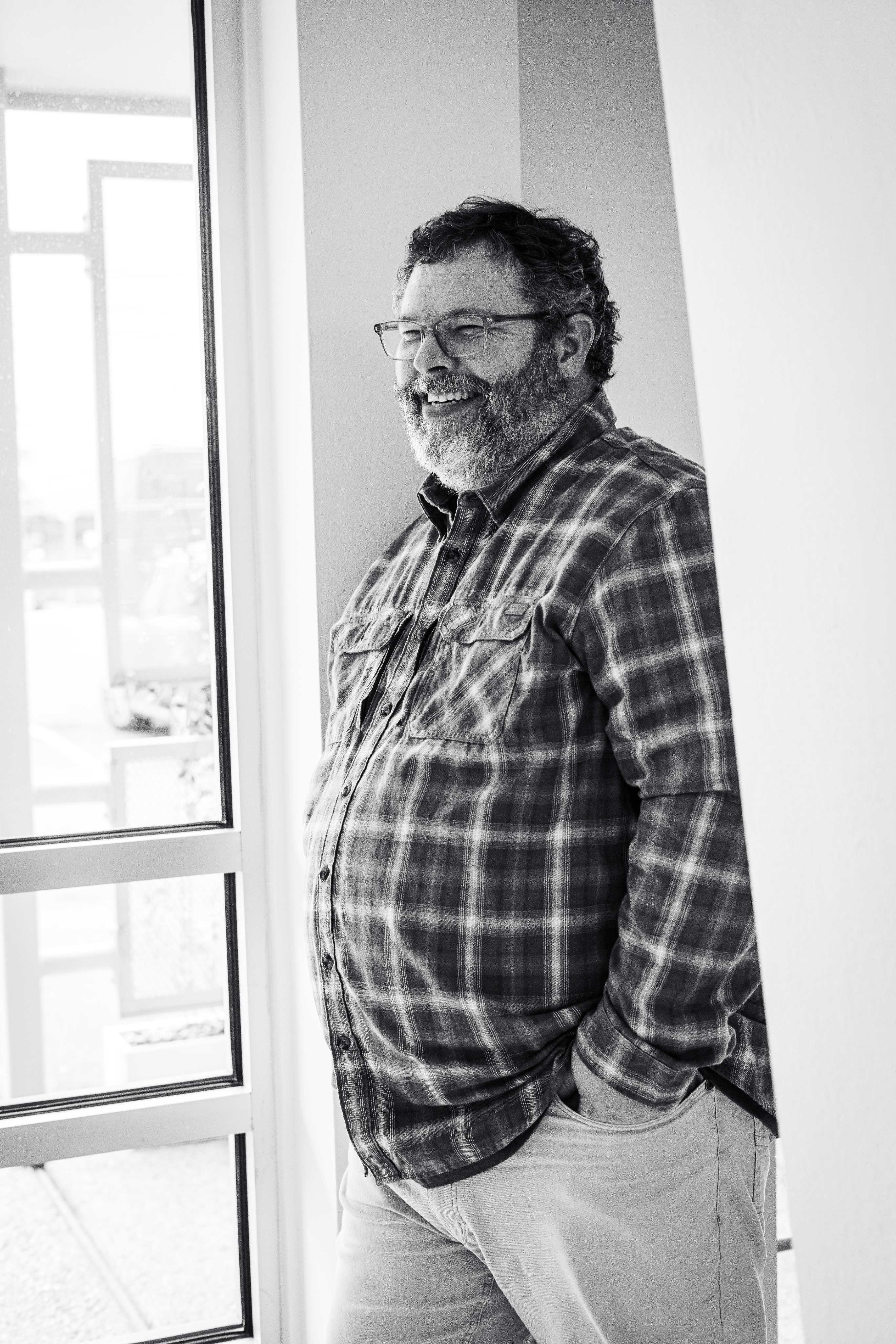 Business Duos A bearded man wearing glasses and a plaid shirt stands by a window indoors, hands casually in his pockets and smiling. This black and white image reflects the relaxed confidence often found in husband-and-wife business partners, showcasing an example of couples in business together. The photo conveys the harmony and partnership that married couples experience as they run a couple-owned business, balancing marriage and entrepreneurship side by side.